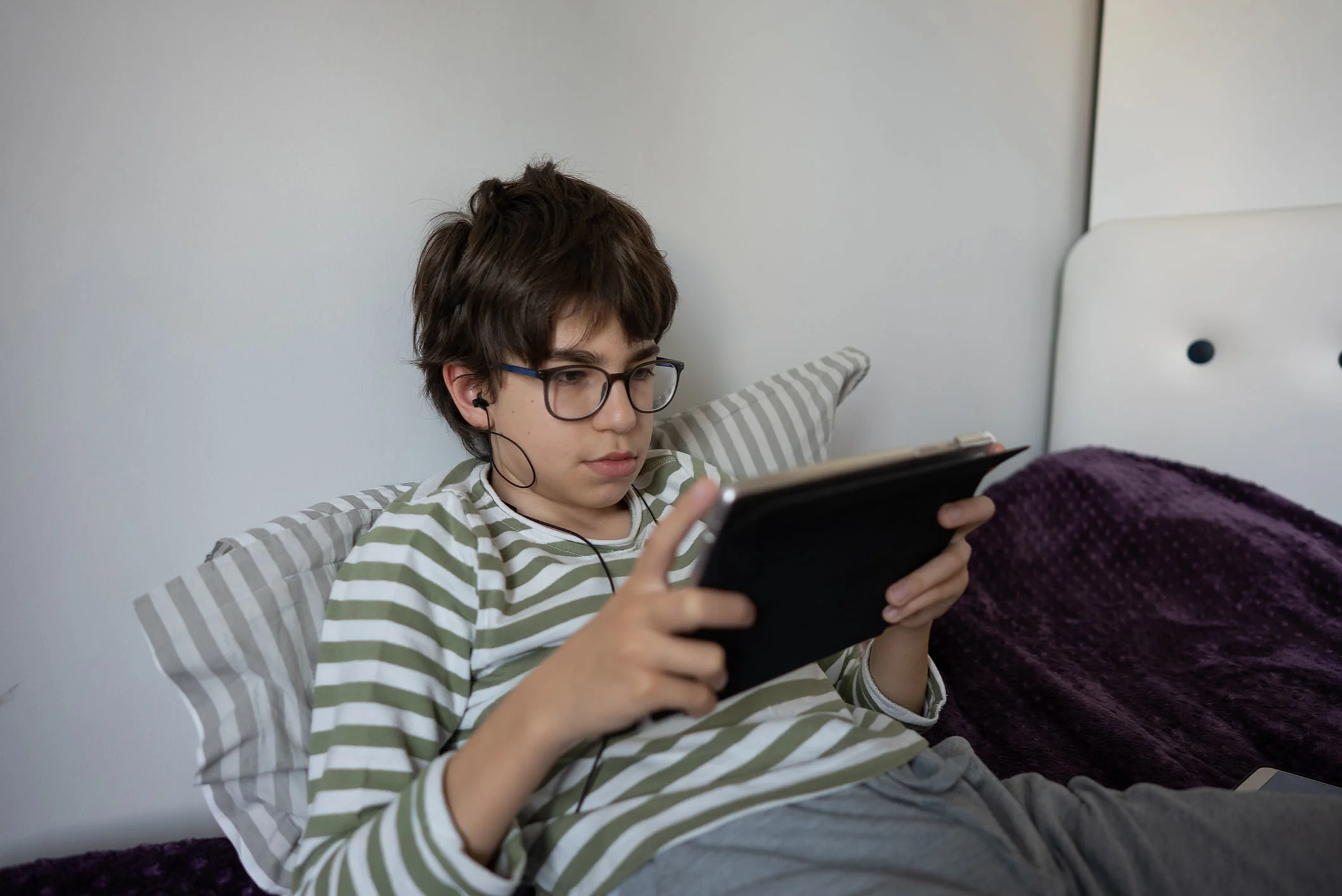 Barn med briller ligger i seng og kigger på iPad med høretelefoner i ørene