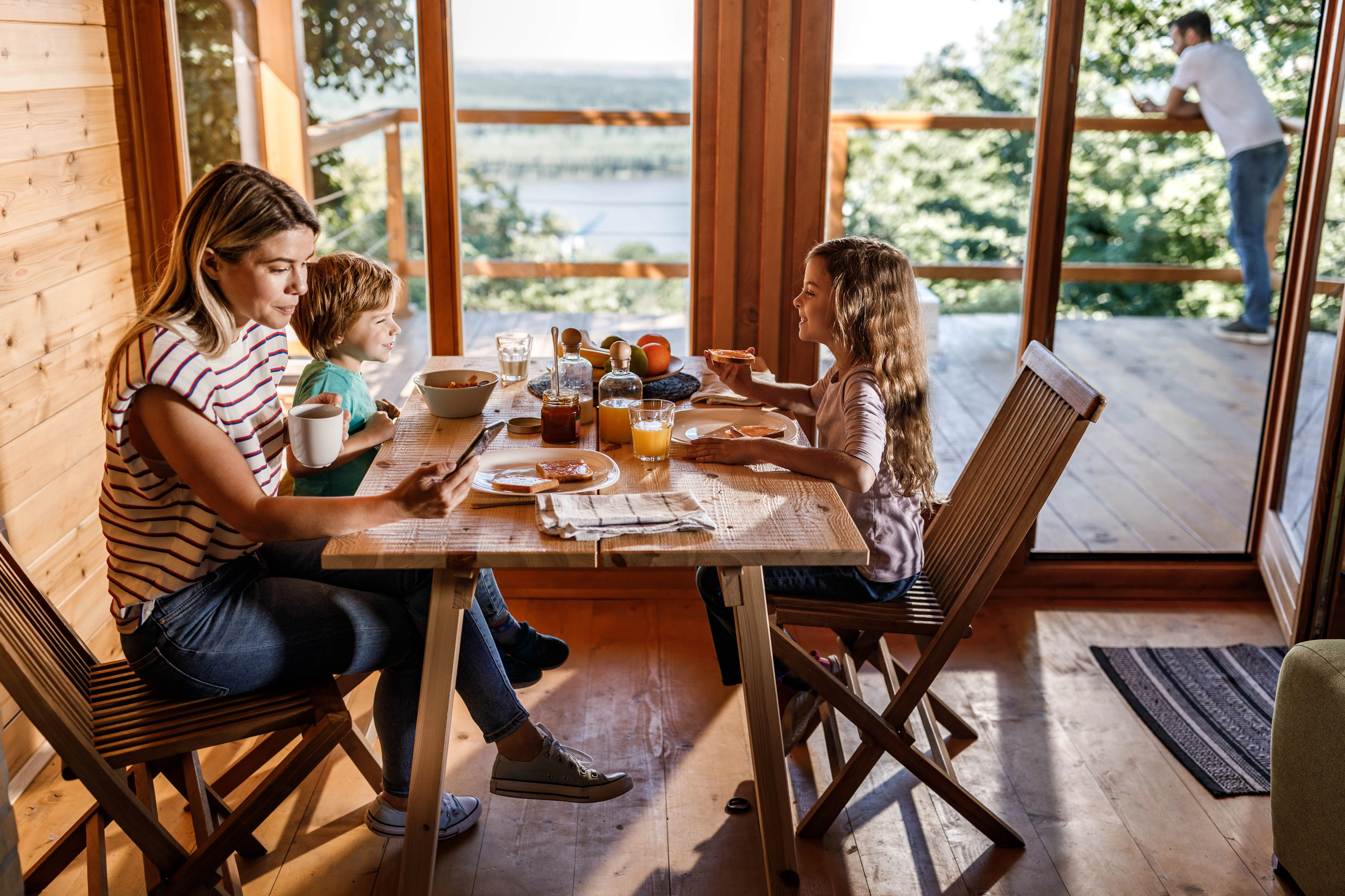 Mor sidder ved spisebord og spiser morgenmad med sine to børn, mens hun kigger på sin smartphone