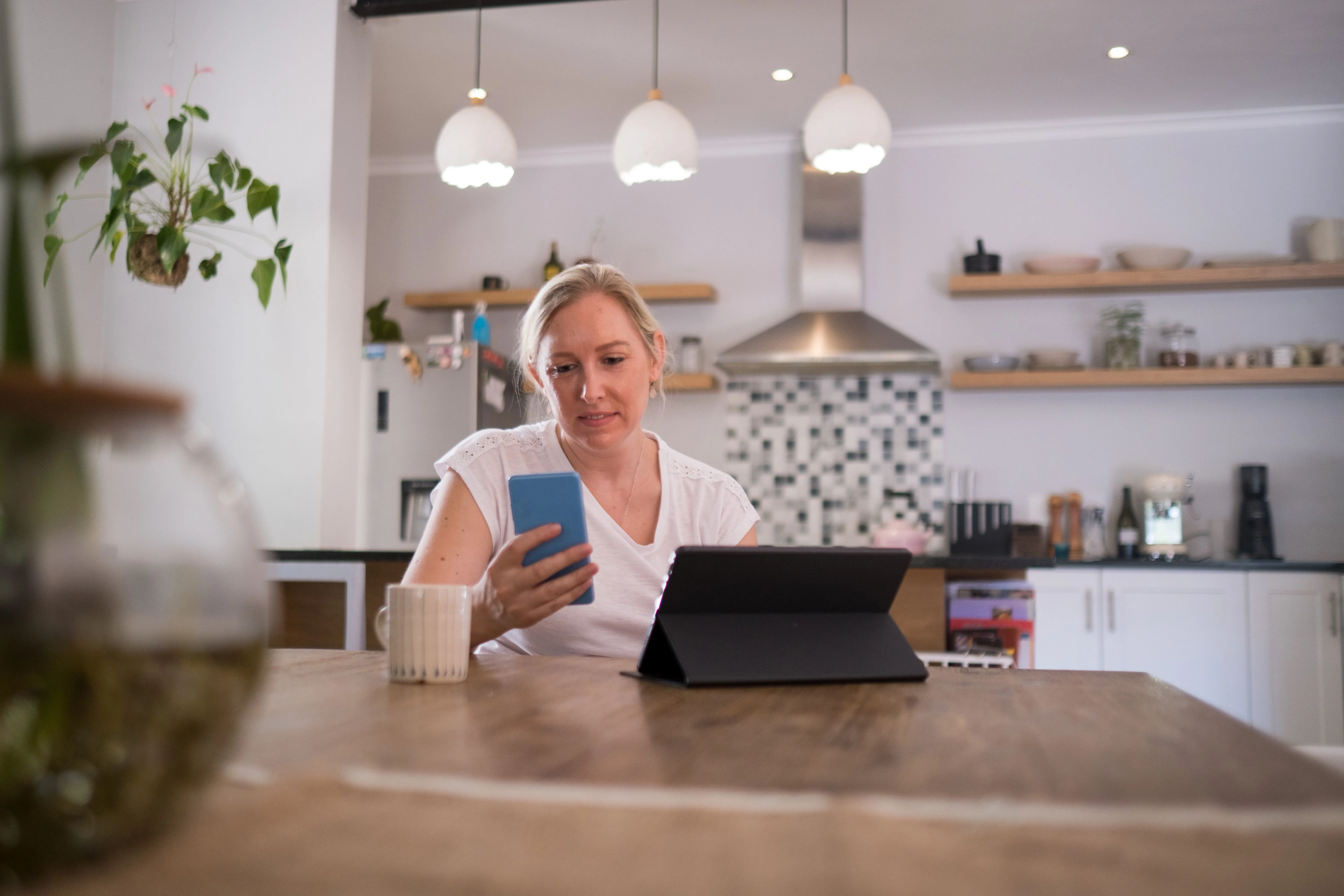 Mor sidder ved spisebord med smartphone i hånden og computer foran sig.