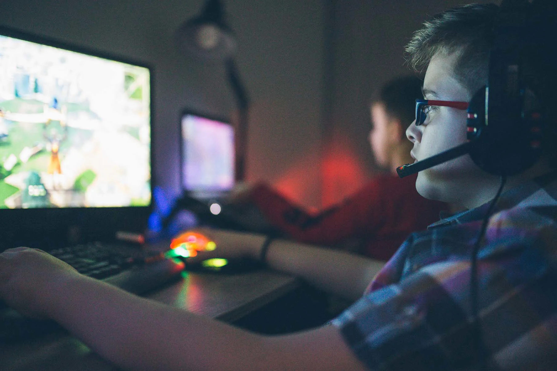 Dreng set fra siden sidder foran computer og gamer med høretelefoner på, mens en anden dreng spiller også i baggrunden.