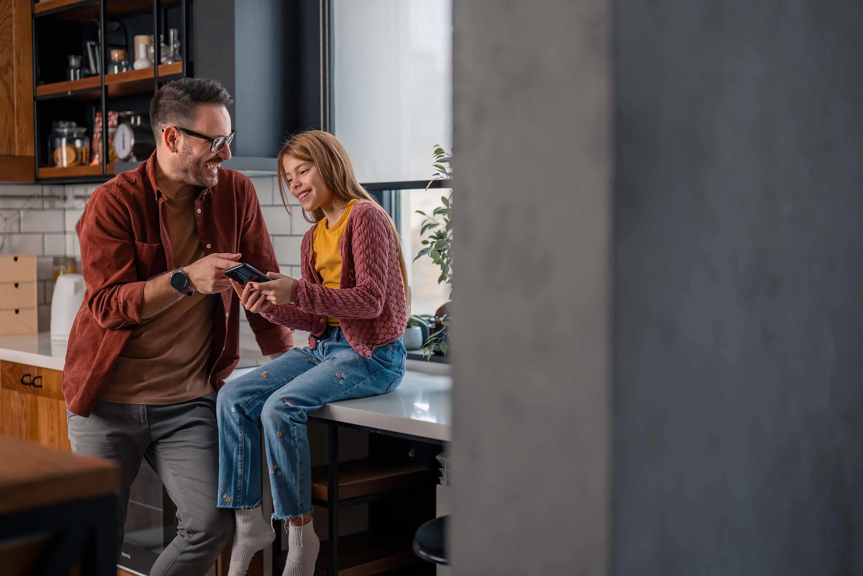 Far og datter står i køkkenet og griner over noget på pigens smartphone