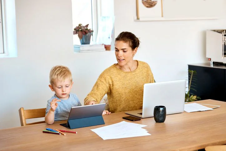 Mor hjælper lille søn med iPad, mens hun sidder og arbejder på computer.