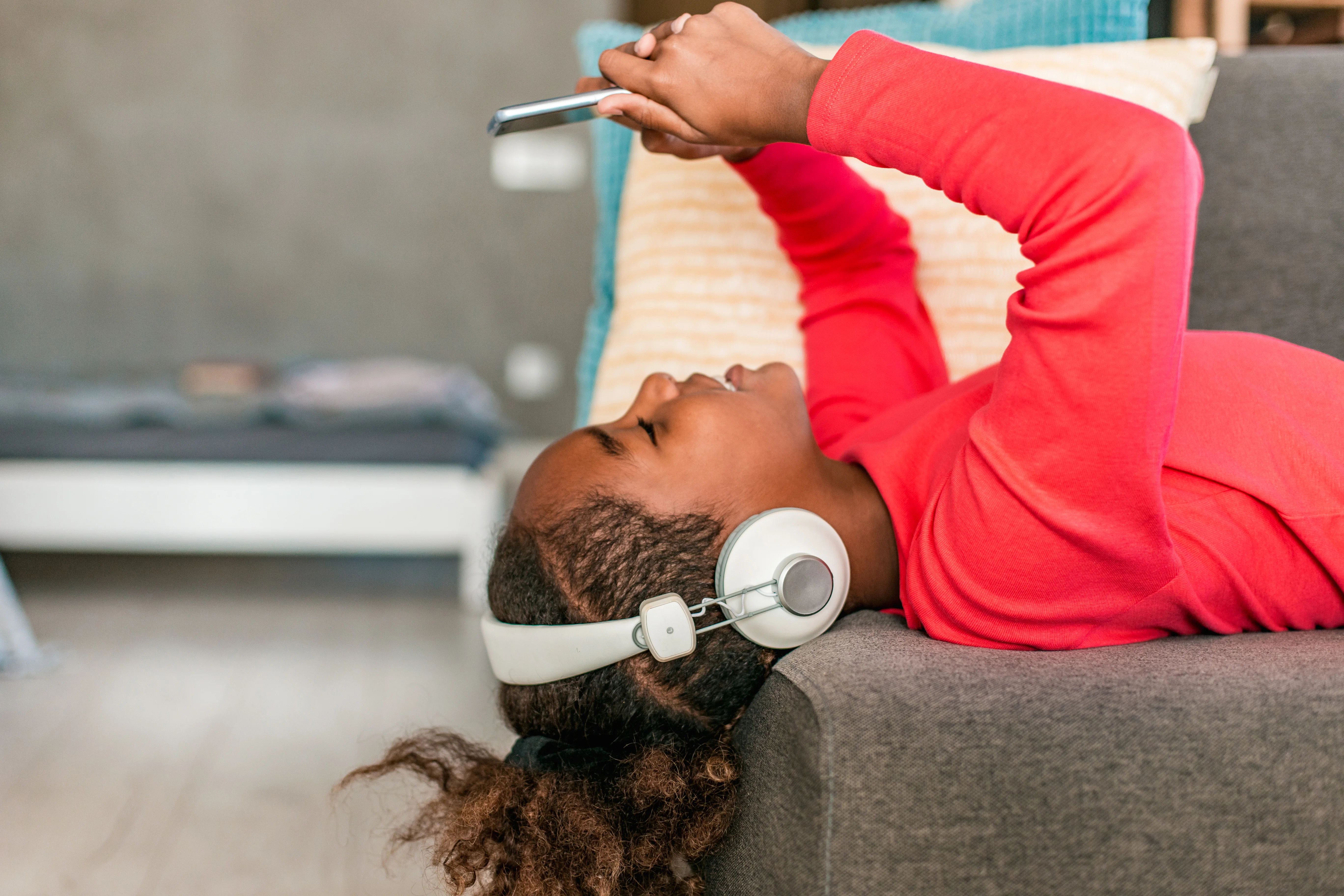 Pige med høretelefoner ligger med hoved ud over sofakanten og ser på sin mobiltelefon