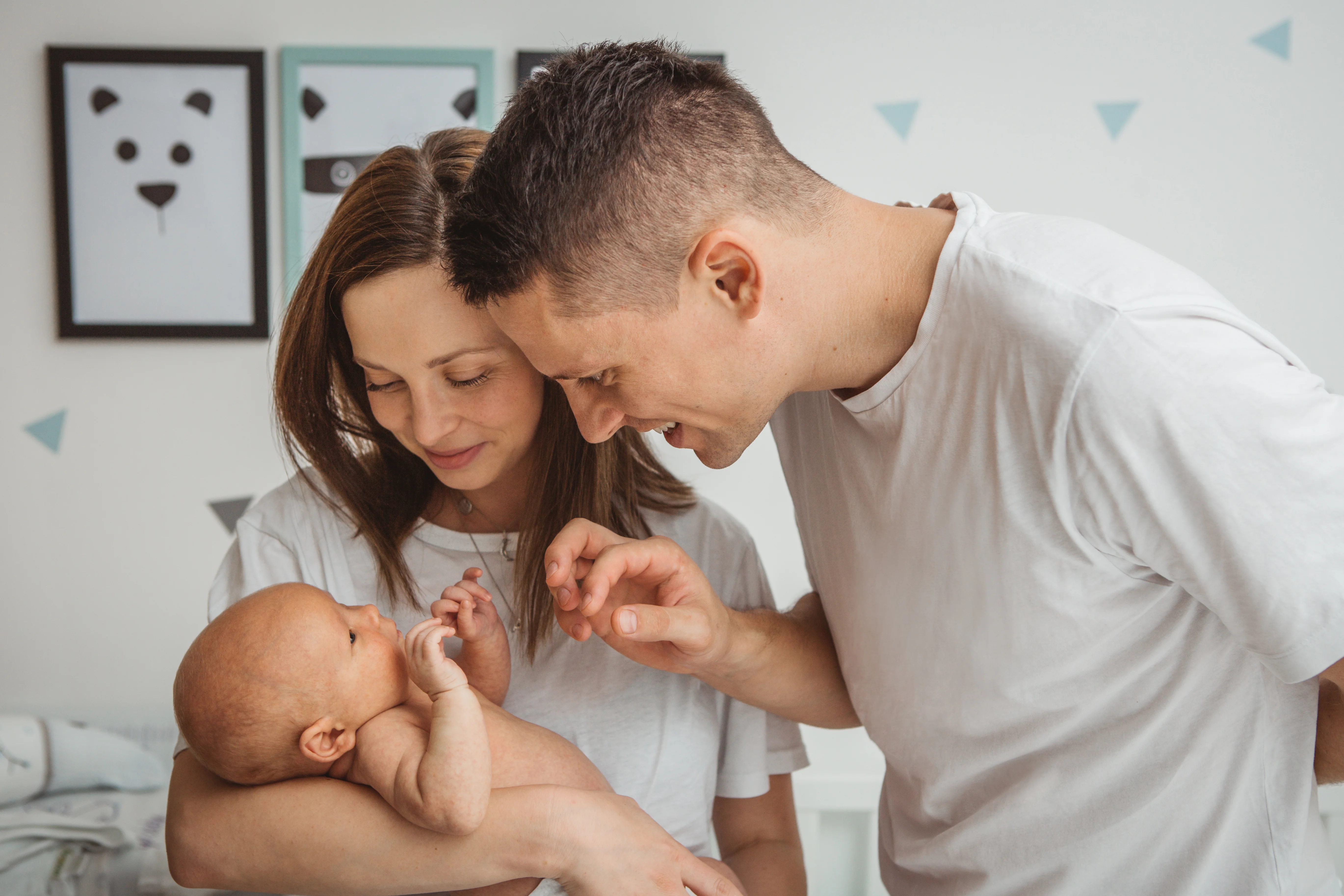 Mor står med lille baby på armen, mens far står ved siden af og skal til at ae baby på maven. Begge kigger på baby.