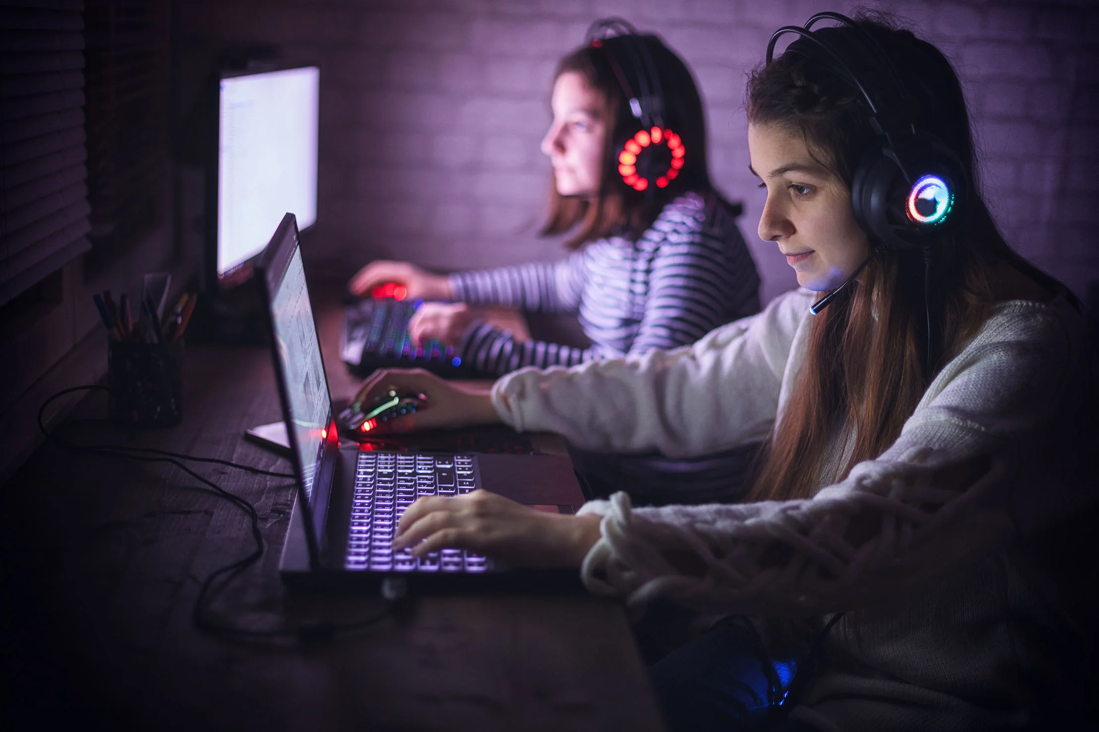 To piger sidder ved skrivebord og gamer med headset på.