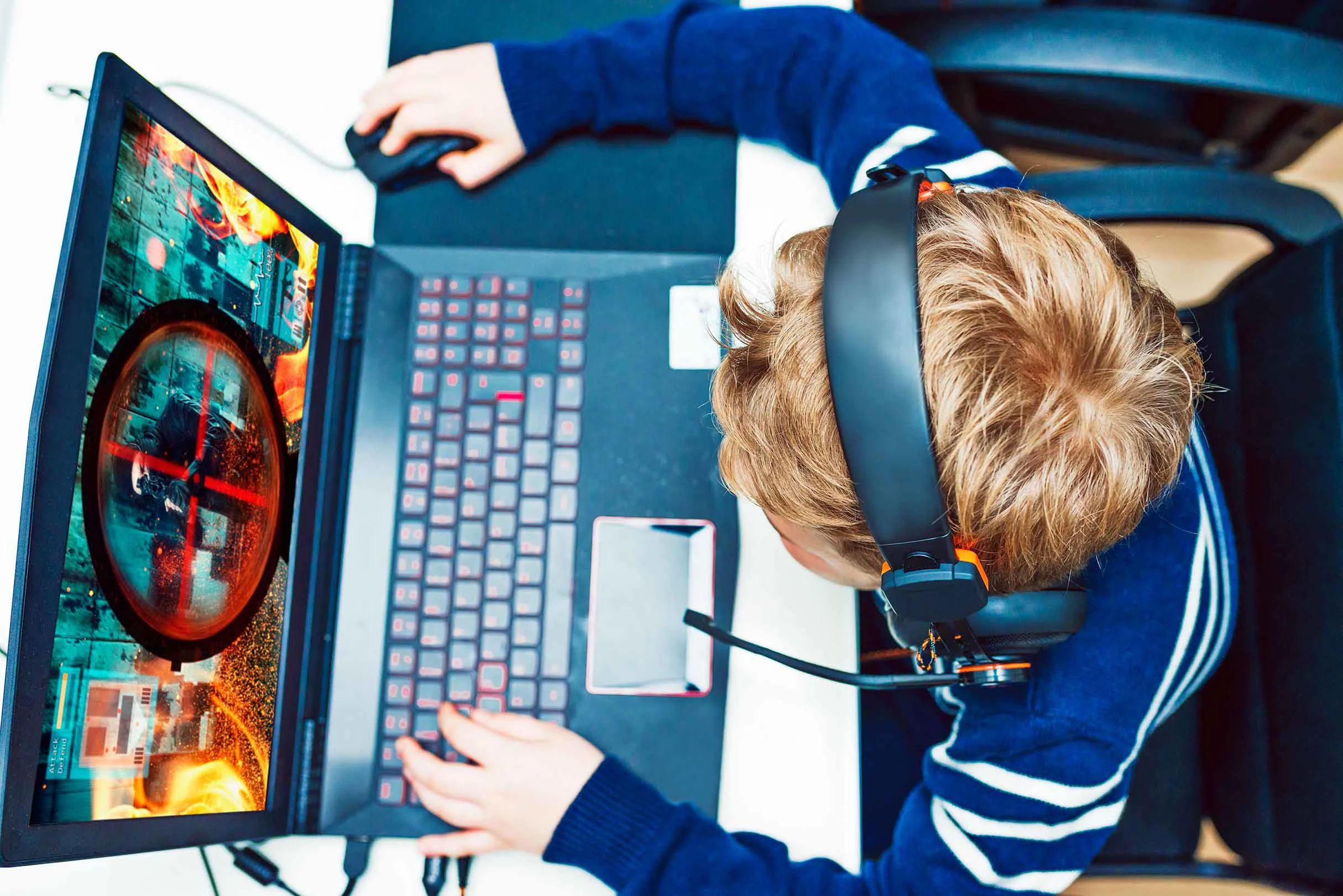 En dreng set oppefra sidder ved skrivebord og spiller computer med høretelefoner på.