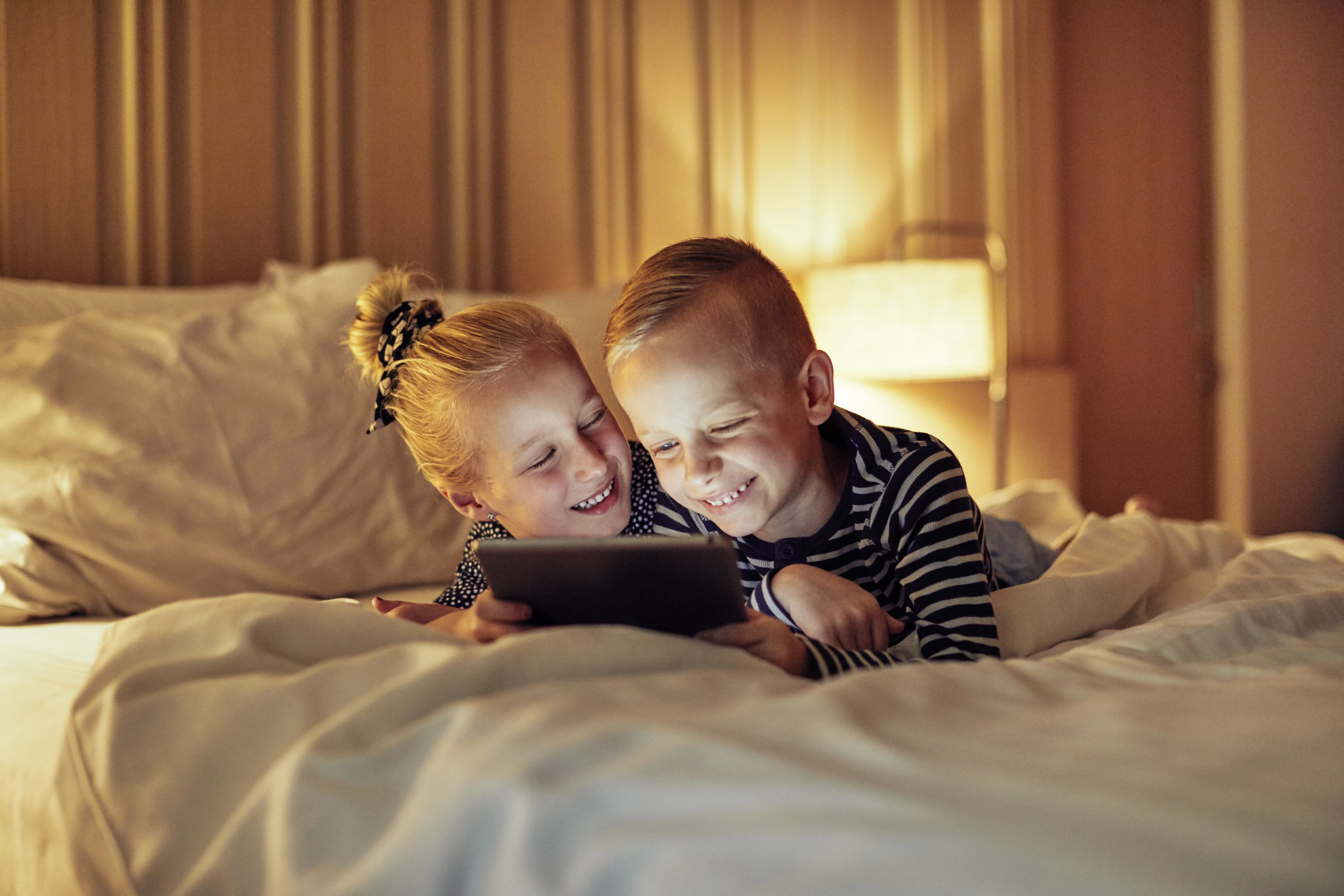 Søskendepar ligger i seng og griner over noget, de ser på deres iPad